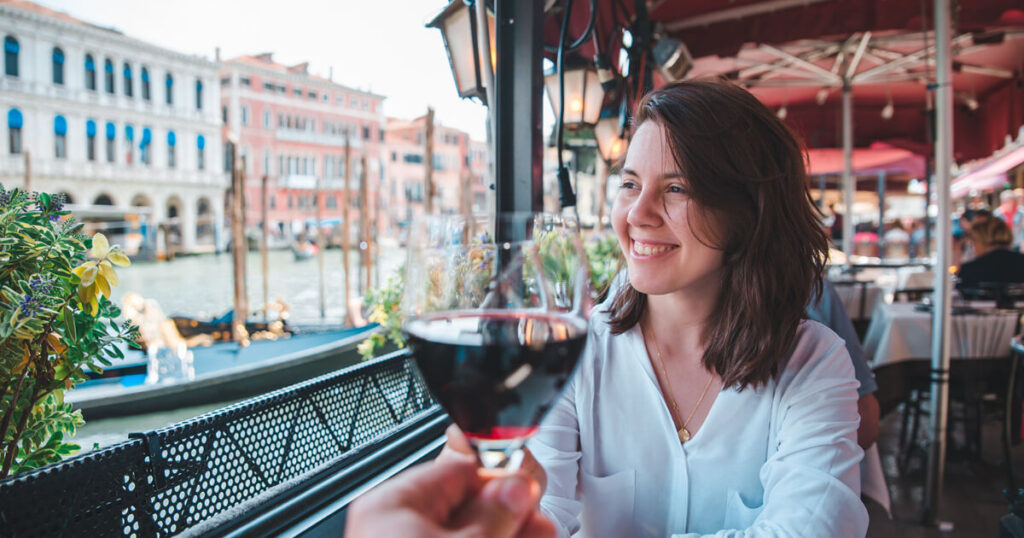 Italian bar and restaurant, boy offering wine to a girl, girl enjoying the view of Venice, gondolas in background, Venice buildings in background.